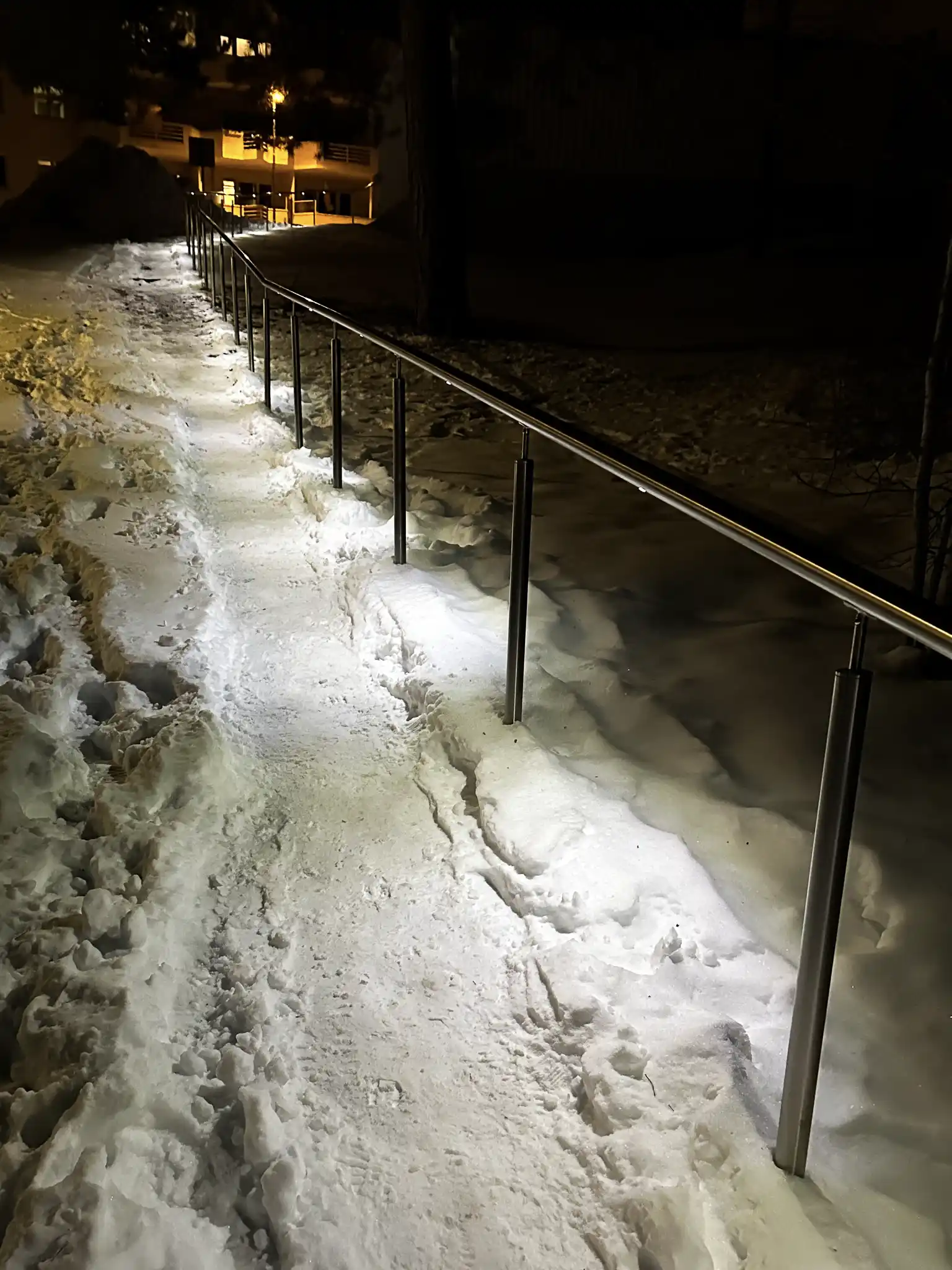 Håndløper på stender med LED-belysning i vintermørket på en snødekt sti..